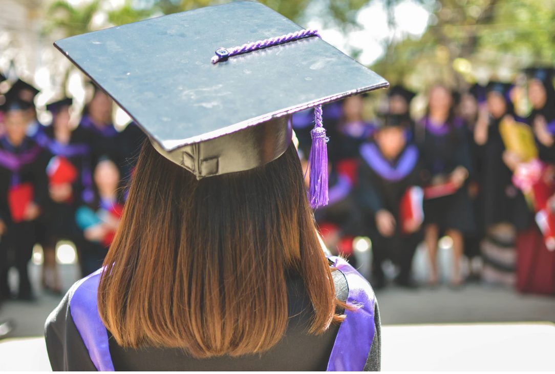 View of graduate's head from the back. They are wearing cap and gown, and other graduates similarly dressed in the background.